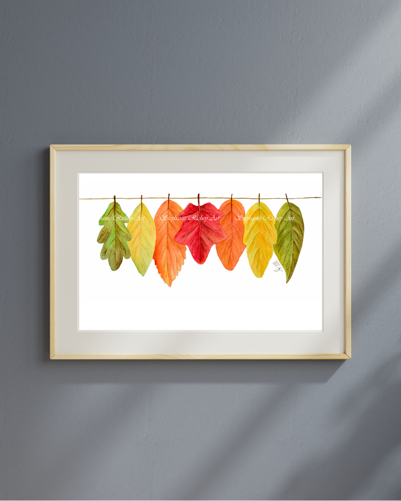 Watercolor artwork of green, yellow, orange and red leaves hanging on a line. Framed in a brown wood frame with a light gray mat. Hanging on a dark gray wall with light filtering in on the right