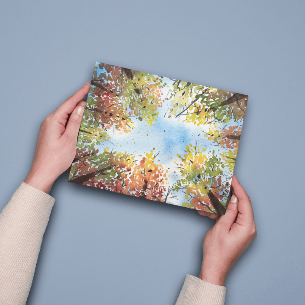 Woman holding a watercolor art print against a gray background. Watercolor print is the perspective of looking up through the fall trees at a cloudy blue sky, with leaves falling down through through the opening.